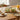 A bright, minimalist dining table set with a set of two green glass dishes with wooden tray, surrounded by neutral-toned plates, fresh bread, orange juice in a glass jug, and a small vase with eucalyptus leaves, softly lit by natural daylight through a nearby window.