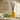 A yellow diffuser bottle with black sticks on a wooden surface with green leaves and a beige wall in the background.